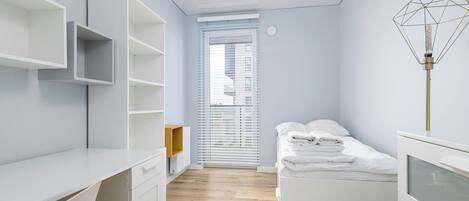 A modern, minimalist bedroom with a wooden floor, white furniture, and a window with blinds.