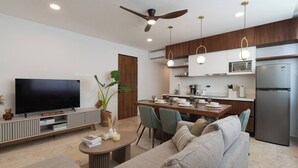 A modern living room with a flat-screen TV, a sofa, a coffee table, and a dining area with chairs and a kitchen in the background.
