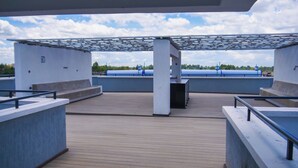 An outdoor terrace with a pergola, concrete seating areas, and a view of a blue water body and greenery in the background.