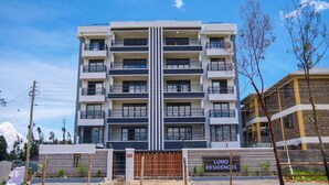 A multi-story residential building with a central vertical feature, balconies with patterned railings, and a gated entrance.