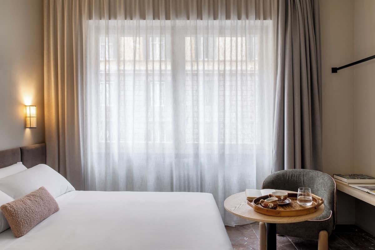 A hotel room with a neatly made bed, a small round table with a tray of food and glasses, and a chair.
