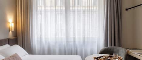 A hotel room with a neatly made bed, a small round table with a tray of food and glasses, and a chair.
