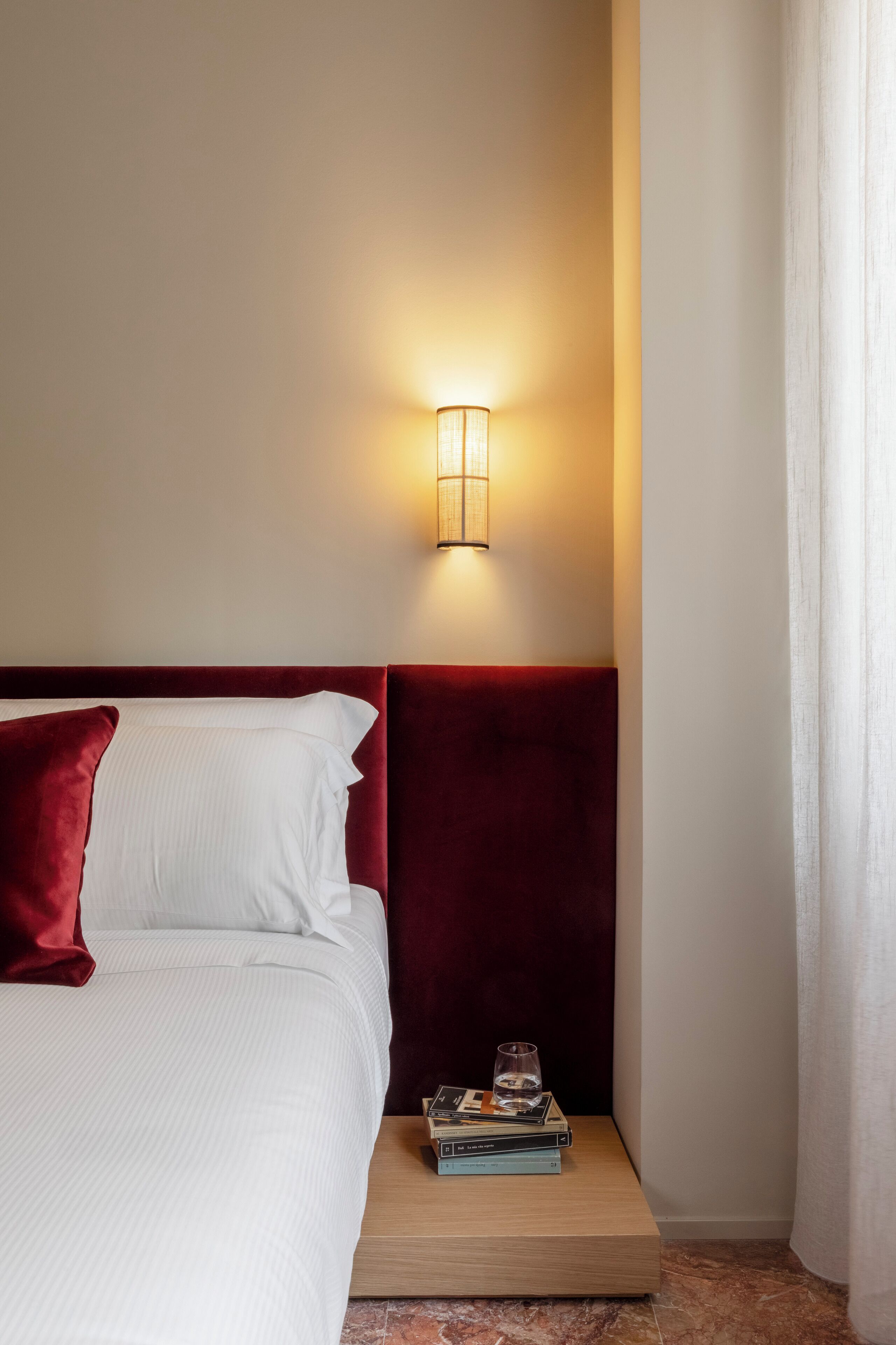 Un lit bien fait, avec du linge de lit blanc et une tête de lit rouge, une table de chevet avec des livres et un verre d’eau, et un luminaire fixé au mur.