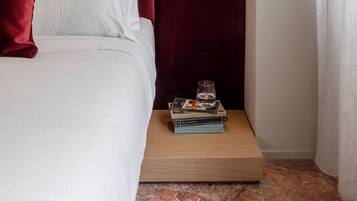 Un lit bien fait, avec du linge de lit blanc et une tête de lit rouge, une table de chevet avec des livres et un verre d’eau, et un luminaire fixé au mur.
