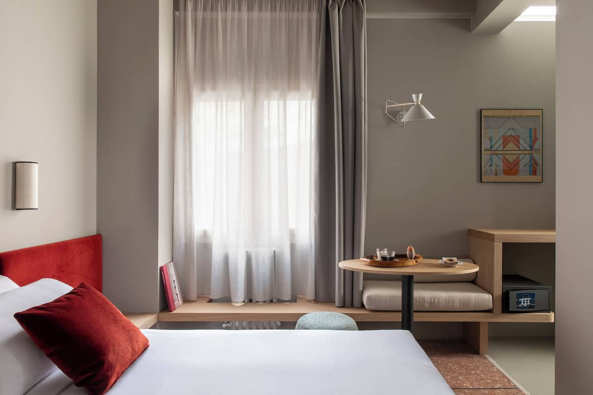 A modern hotel room with a bed, a bedside table, a small round table with a tray, a wall-mounted lamp, and a painting.