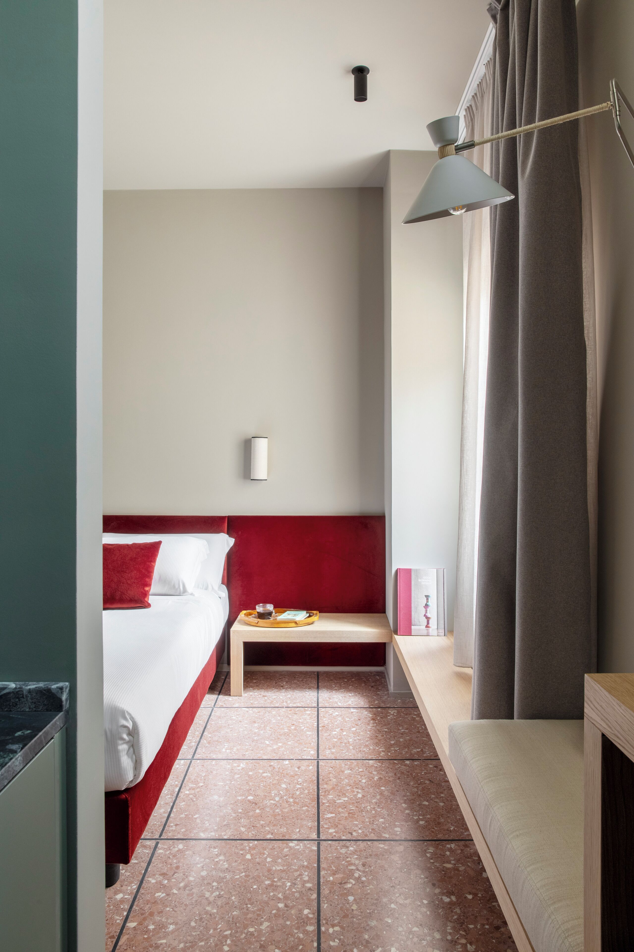 Habitación de hotel con cama, mesita de noche, una pequeña mesa con bandeja, luz en la pared y ventana con cortinas.
