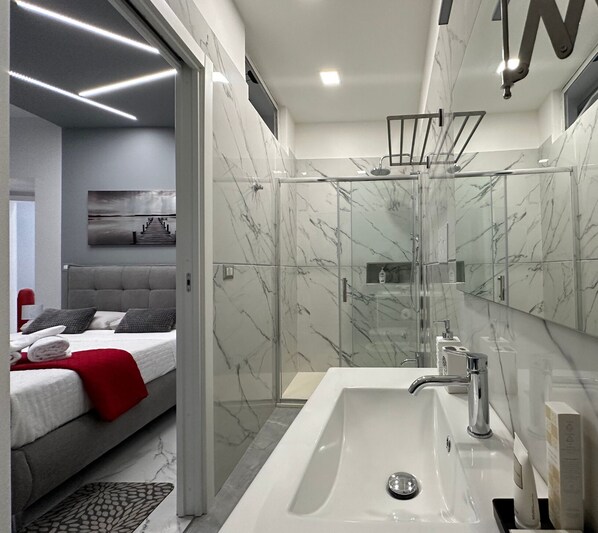 A modern bathroom with a glass-enclosed shower, a large white sink, and a mirror with a marble backsplash. - Sweet Tide Suites (Licata)