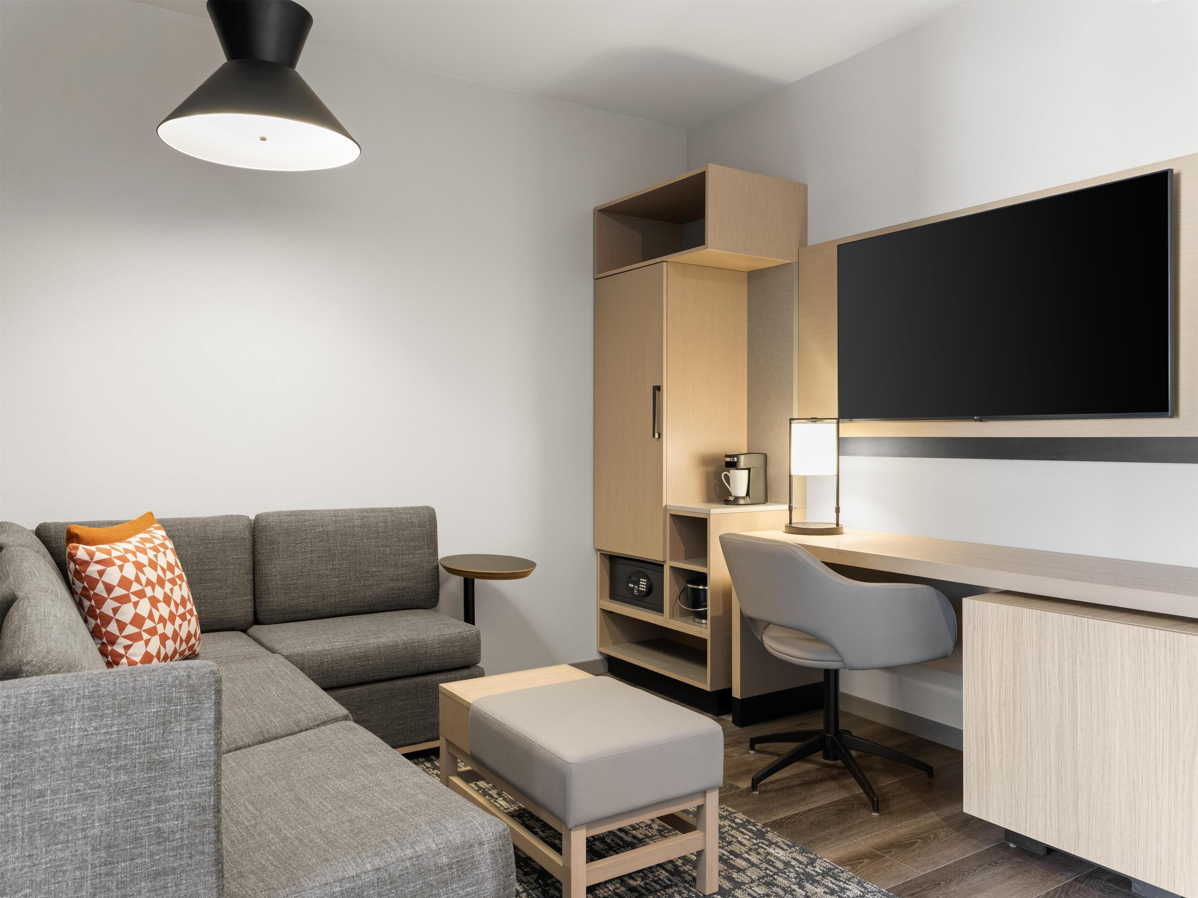 Ein modernes Hotelzimmer mit grauem Sofa, einem kleinen runden Tisch, einem Schreibtisch mit Stuhl, einer Kaffeemaschine und einem großen Flachbildfernseher an der Wand.