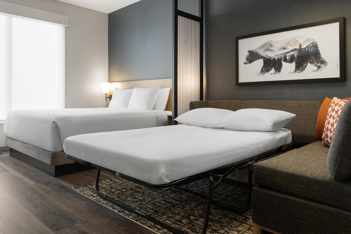 A hotel room with two beds, a sofa, a rug, and a mountain landscape painting on the wall.