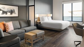 Habitación de hotel moderna con una cama grande, un sofá gris, una pequeña mesa de madera, un escritorio y un amplio ventanal con vista al mar.