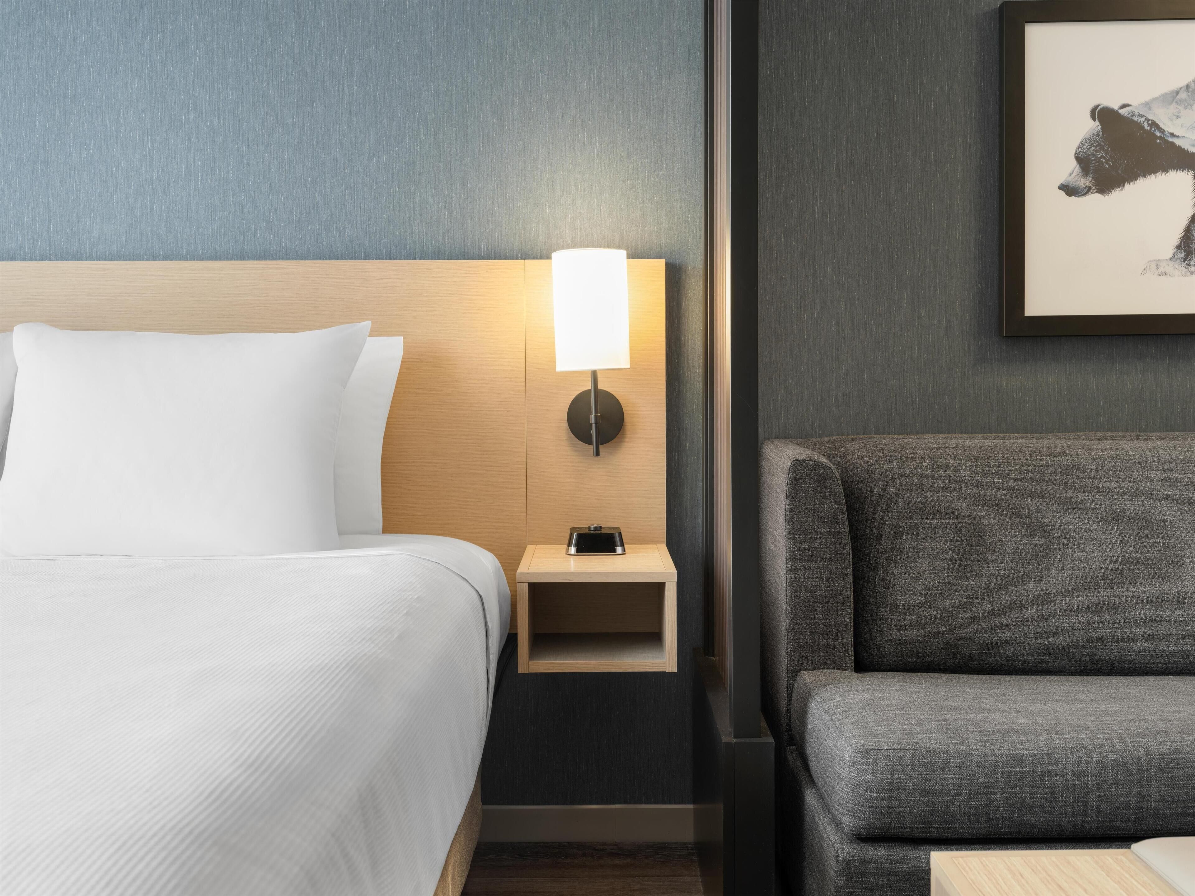 A hotel room with a bed, a nightstand with a lamp, a gray armchair, and a framed picture on the wall.