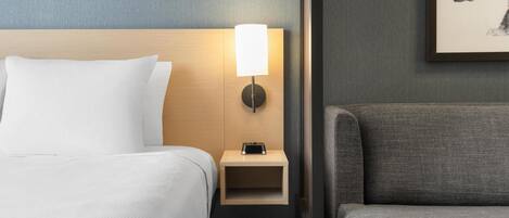 A hotel room with a bed, a nightstand with a lamp, a gray armchair, and a framed picture on the wall.