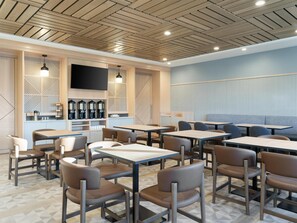 A modern cafe with wooden ceiling panels, multiple tables and chairs, a coffee station, and a television screen.