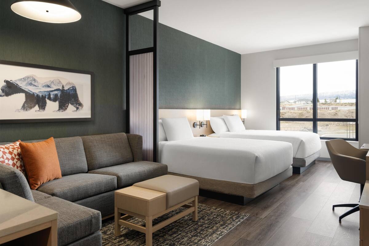 A modern hotel room with a gray sofa, two beds, a desk, and a large window offering a view of the mountains.