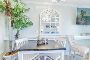 A dining area with a wooden table, white chairs, a large window with a decorative grille, and a potted plant.