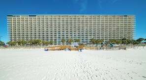 A large hotel with multiple floors and balconies, located on a sandy beach with palm trees and a playground.
