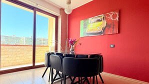 A dining area with a red wall, a painting of a car, and a view of buildings through a large window. - Spacious 2BR Apartment for 6 • Parking • 20 min Walk to Beach (Quartu Sant'Elena)