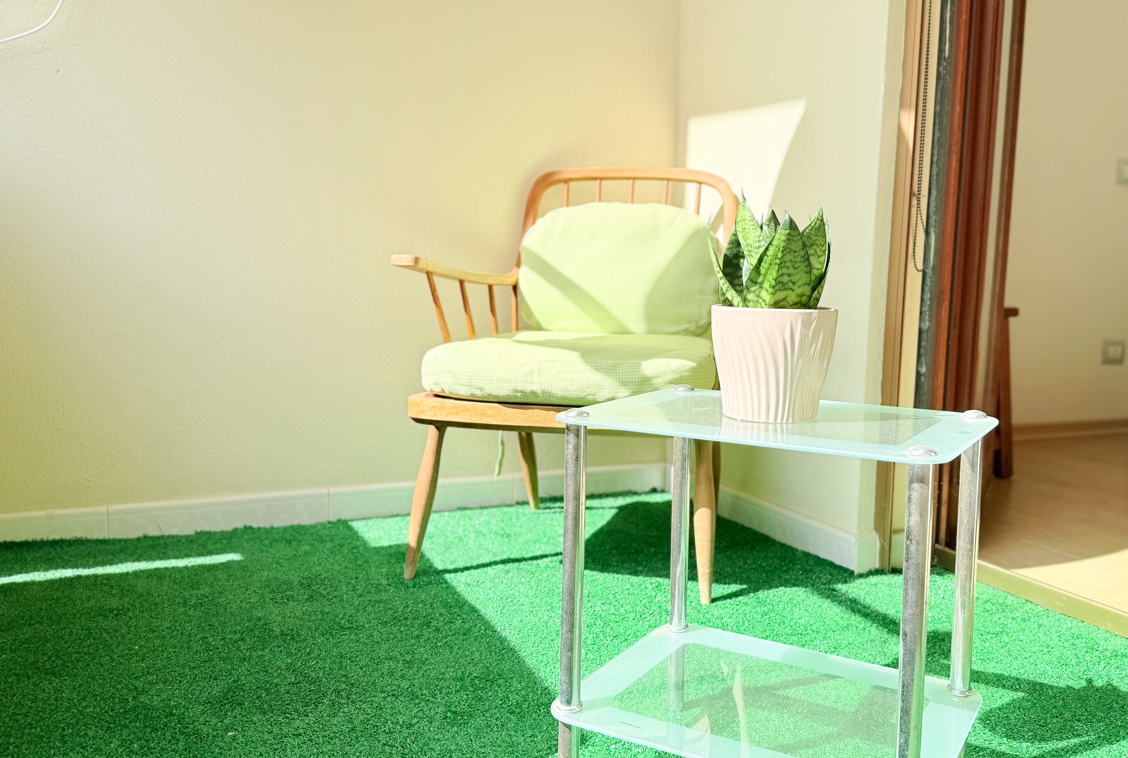 Una habitación con suelo de césped artificial verde, una silla de madera con cojín y una mesa de vidrio con planta en maceta.