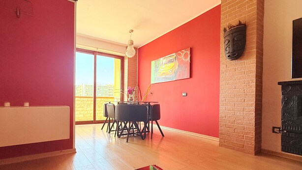 A dining area with a table and chairs, a red wall with a painting, and a brick fireplace. - Spacious 2BR Apartment for 6 • Parking • 20 min Walk to Beach (Quartu Sant'Elena)