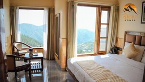A hotel room with a bed, a desk, and a chair. There are curtains on the windows, and a view of mountains is visible. - Dogri Homes (Shimla)