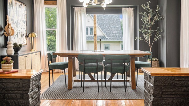 A dining area with a wooden table and four chairs, a black and white rug, and a view of a house through large windows.