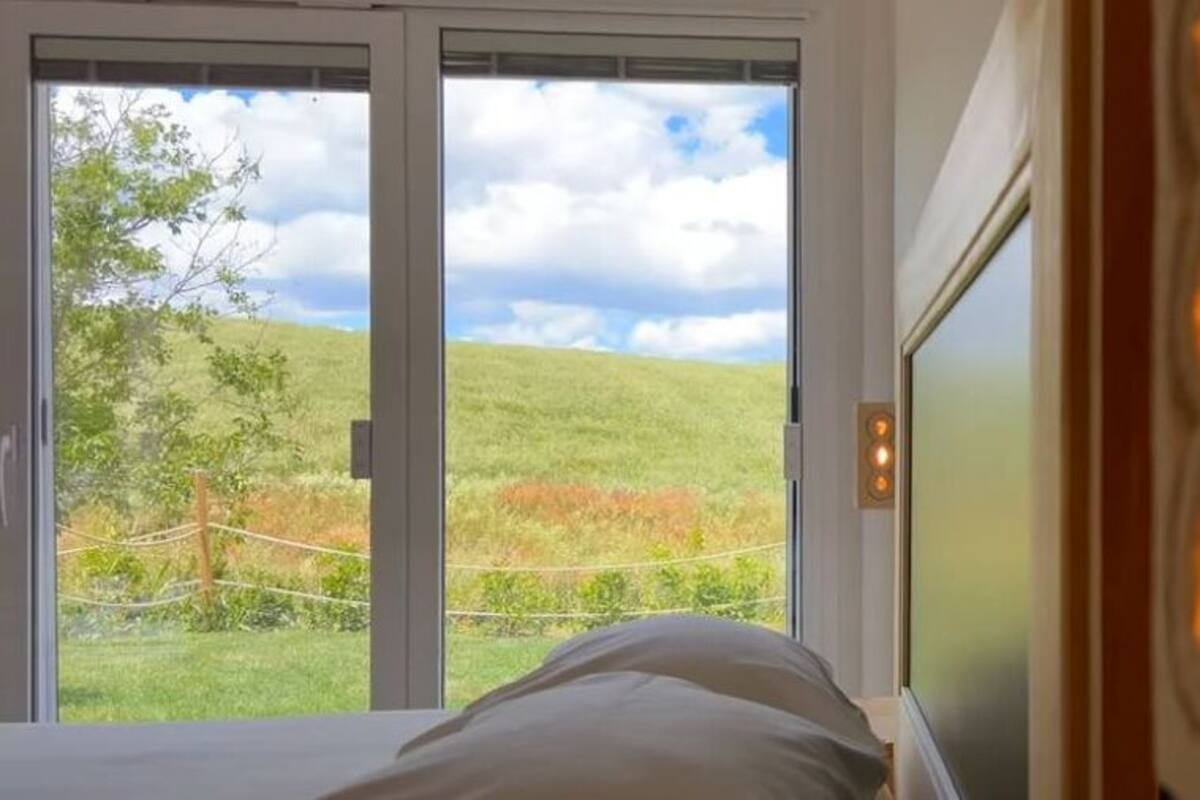 A bedroom with a large window, a bed, and a view of a grassy landscape.