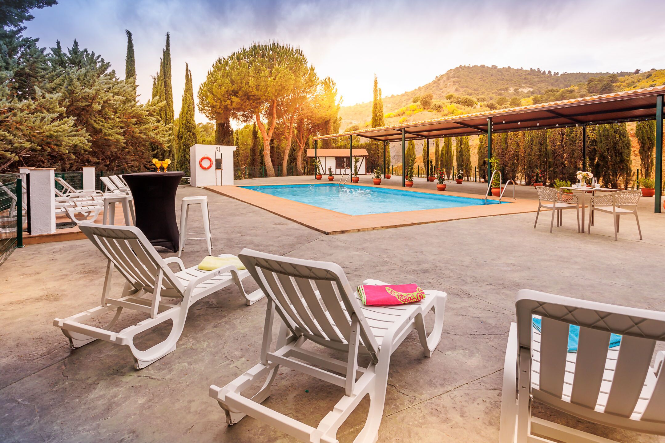 Un espace aménagé au bord de la piscine, avec des chaises longues, un coin salon abrité et une vue sur les arbres et les collines.