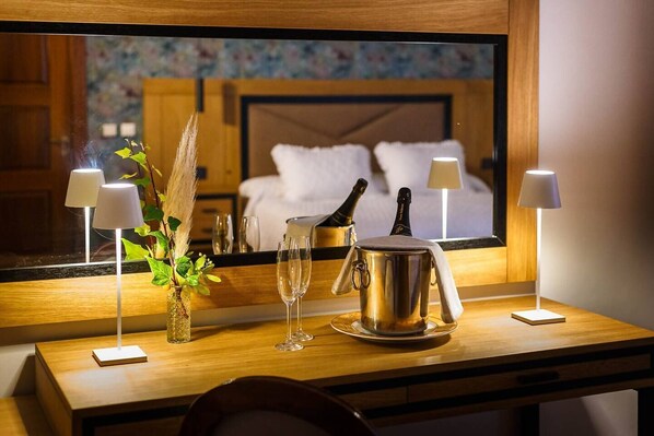 A hotel room with a wooden desk, two bedside lamps, a vase with greenery, a champagne bottle, and a metal pitcher.