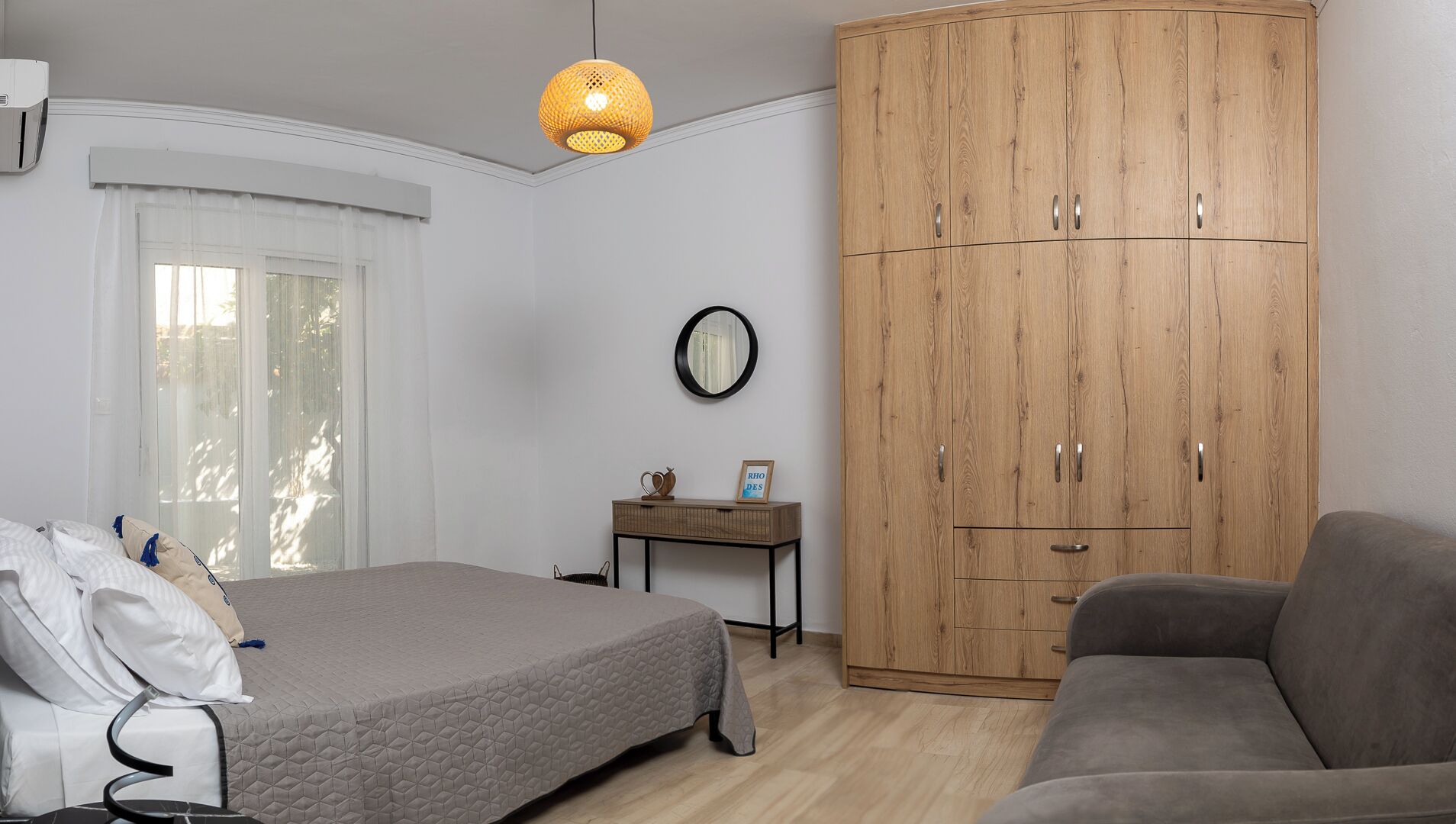 A bedroom with a bed, a grey sofa, a wooden wardrobe, a bedside table with a mirror, and a hanging lamp.
