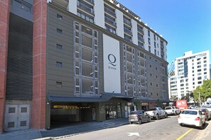 Um edifício com vários andares, um logotipo 'Q' em destaque, uma área de estacionamento com vários carros e um céu limpo.