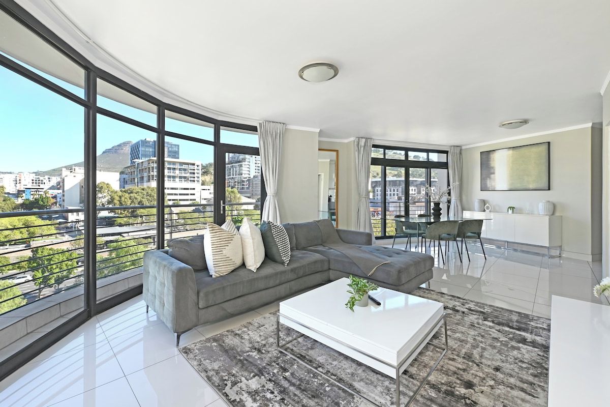 A modern living room with a grey sectional sofa, a white coffee table, and a view of the city through large windows.
