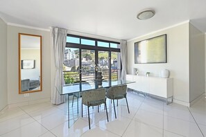 Ein modernes Wohnzimmer mit einem Glastisch, zwei Stühlen, einem großen Fenster mit Vorhängen, einem weißen Schrank und einem gerahmten Kunstwerk an der Wand.