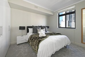 Ein modernes Schlafzimmer mit einem großen Bett, Nachttischen, einem Fenster mit Jalousien und Blick auf Gebäude.