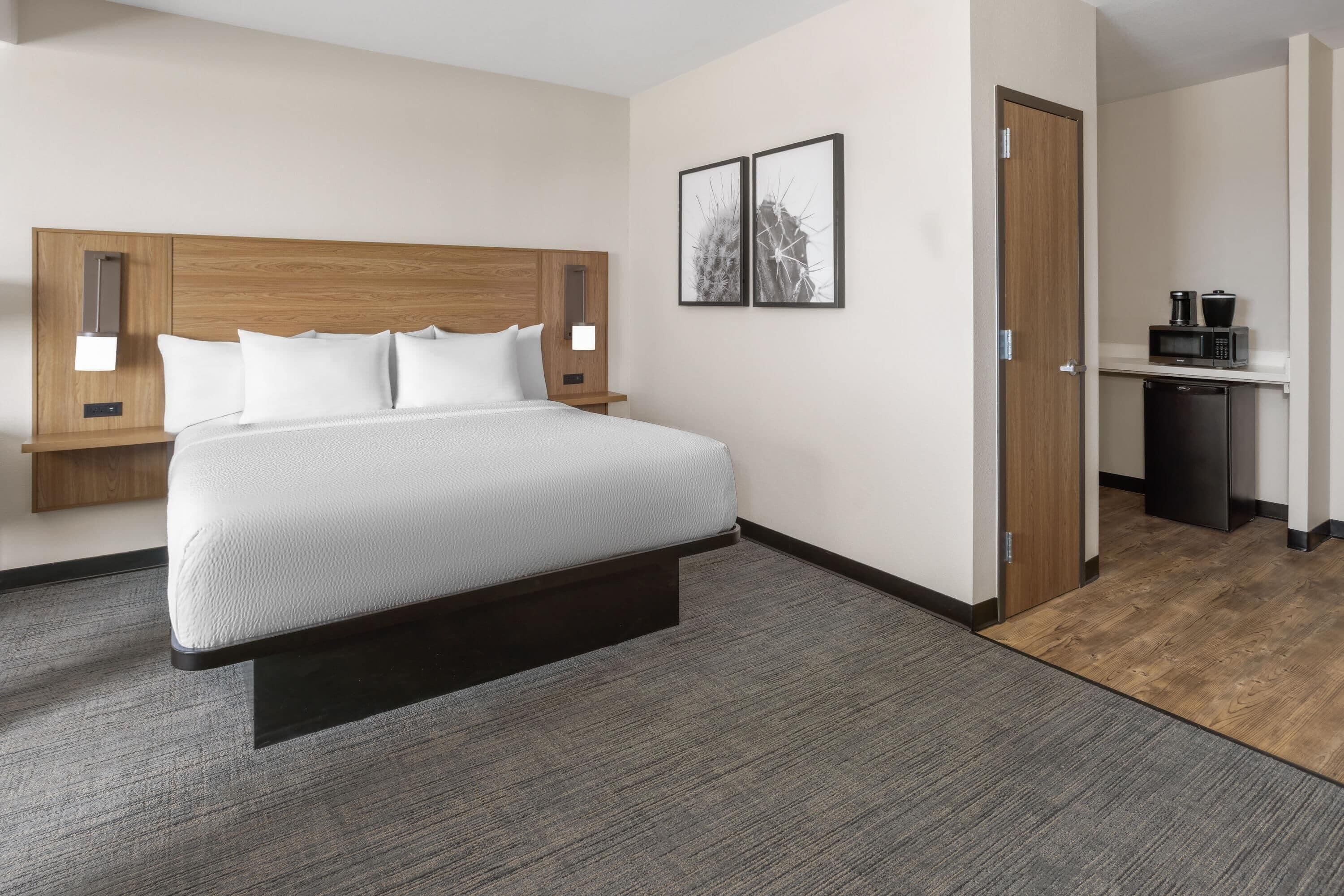 Quarto de hotel com cama, mesas de cabeceira, luz acoplada à parede, porta e área de cozinha compacta.