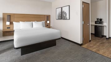 Quarto de hotel com cama, mesas de cabeceira, luz acoplada à parede, porta e área de cozinha compacta.