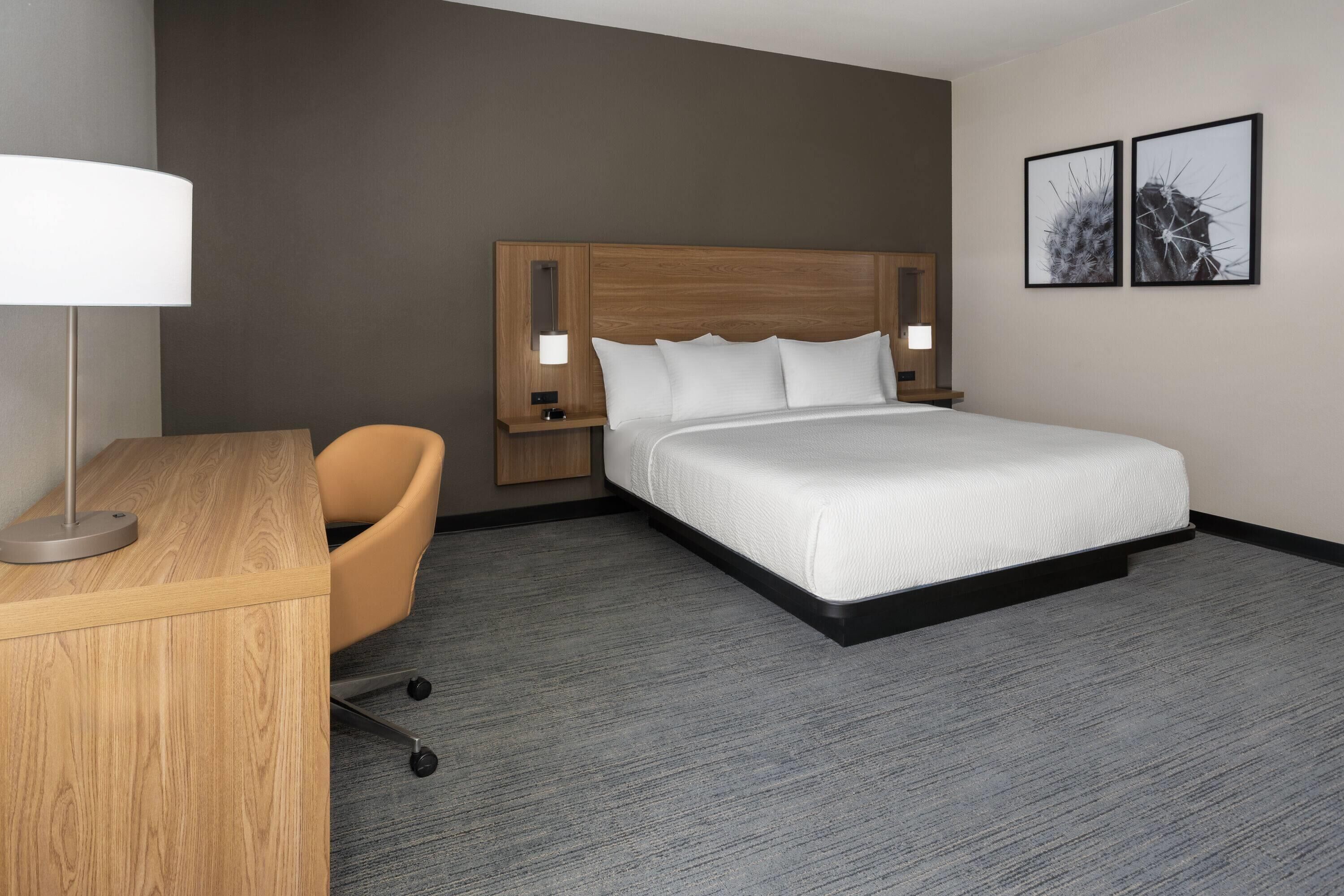 A hotel room with a wooden desk, a single chair, a large bed with a wooden headboard, two bedside lamps, and two framed pictures on the wall.