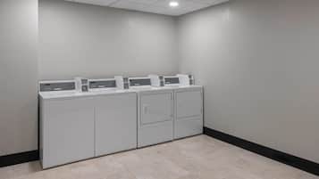 Una sala de lavandería con cuatro lavadoras y secadoras, suelo de baldosas y una pared sencilla con zócalo negro.