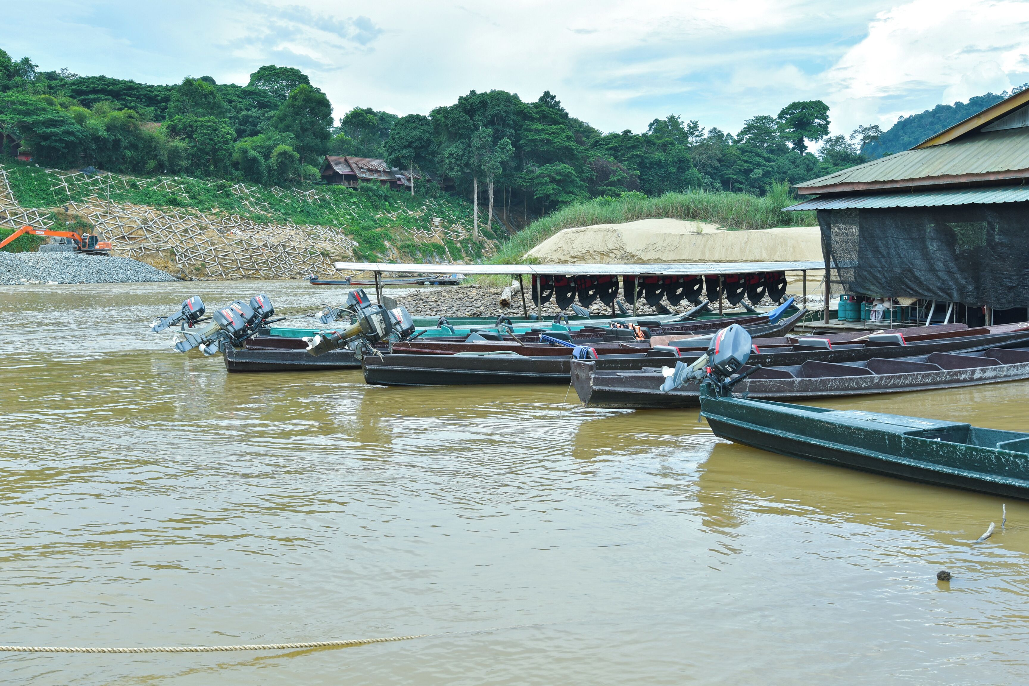 A river with several boats docked, a construction site in the background, and a building with a thatched roof.