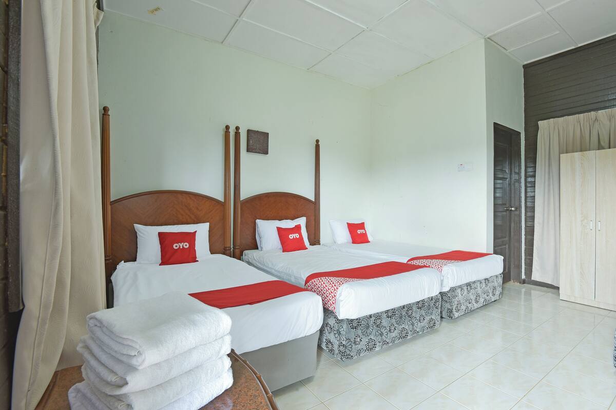 Quarto de hotel com duas camas individuais, cada uma com cabeceira de madeira, roupa de cama branca e almofadas vermelhas. Há uma pilha de toalhas brancas sobre uma mesa de madeira.