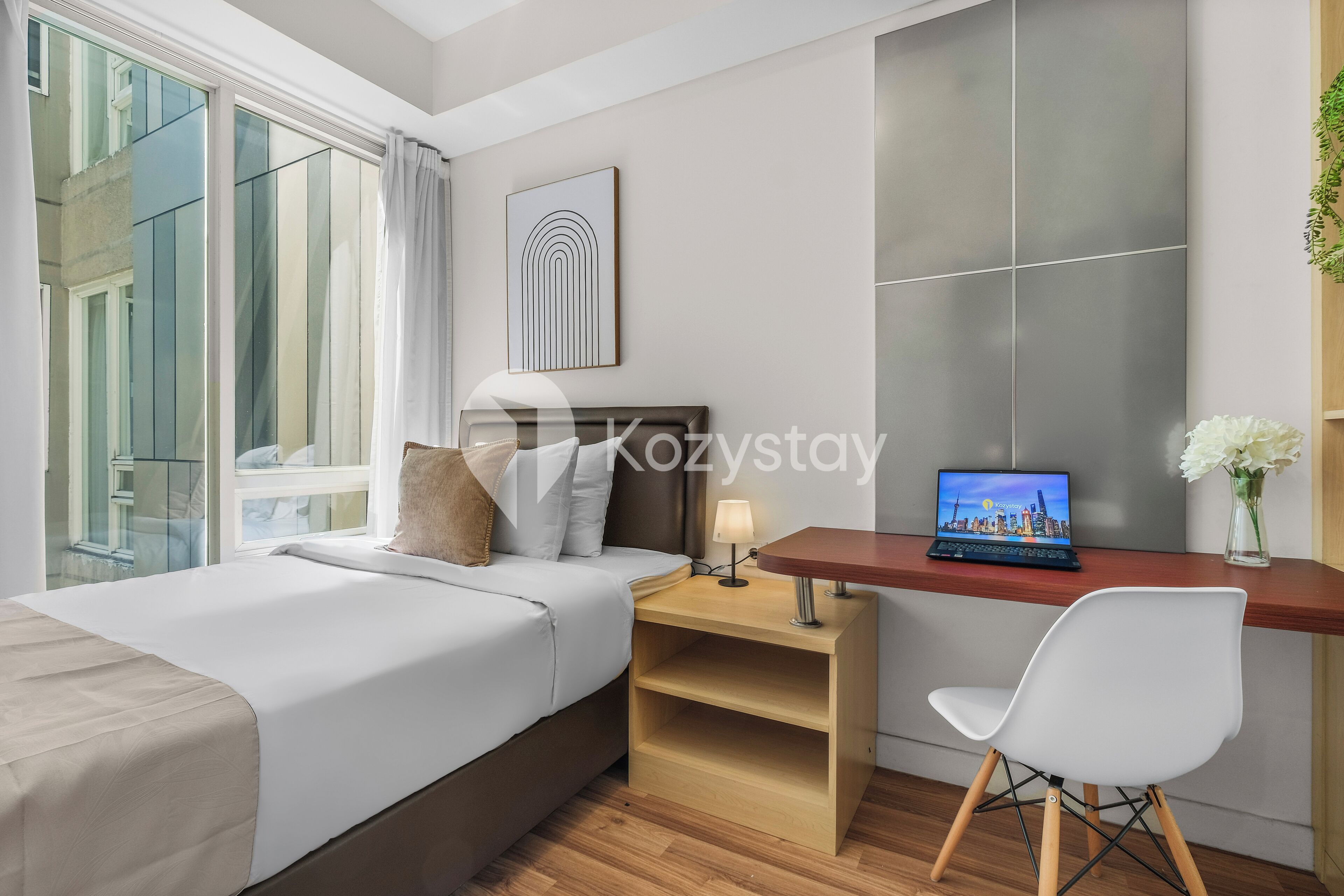 A hotel room with a bed, a desk with a laptop, a chair, and a window with curtains.