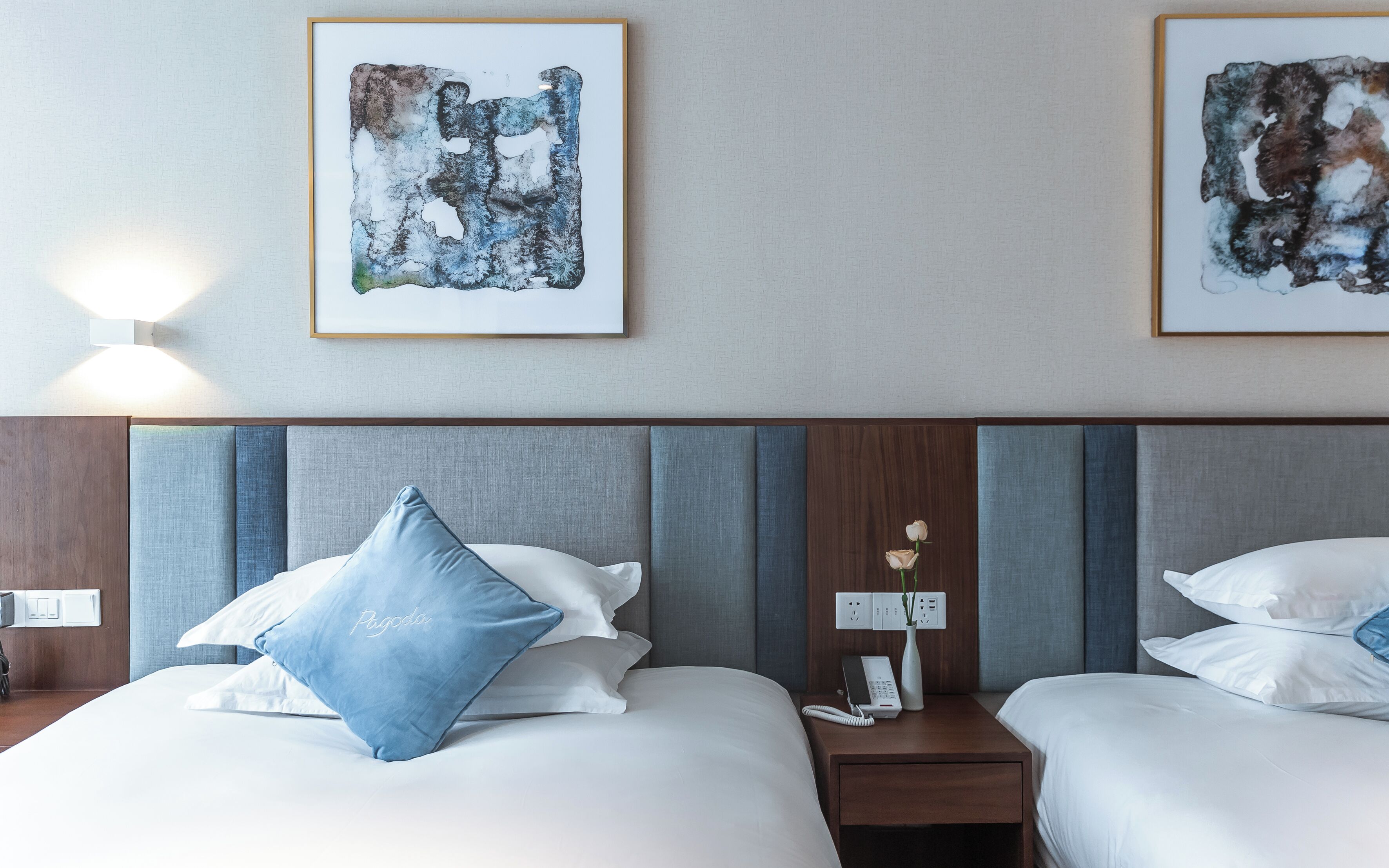 A hotel room with two beds, each with white bedding and blue pillows, a wooden headboard, and abstract artwork on the walls.