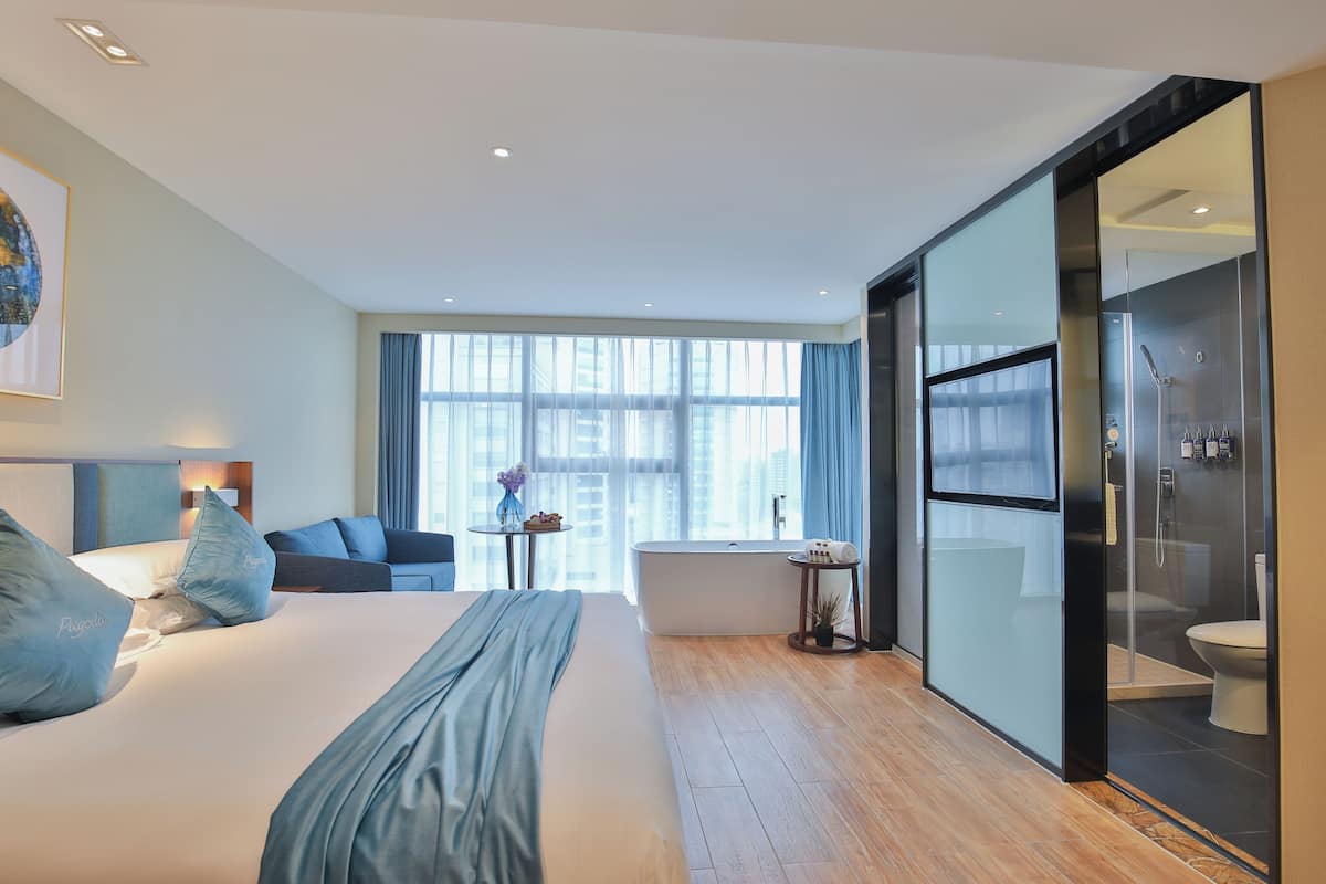 Ein modernes Hotelzimmer mit einem großen Bett, einer Sitzecke mit einer Couch, einem kleinen Tisch mit einer Vase, einer Badewanne und einem gläsernen Duschbereich.