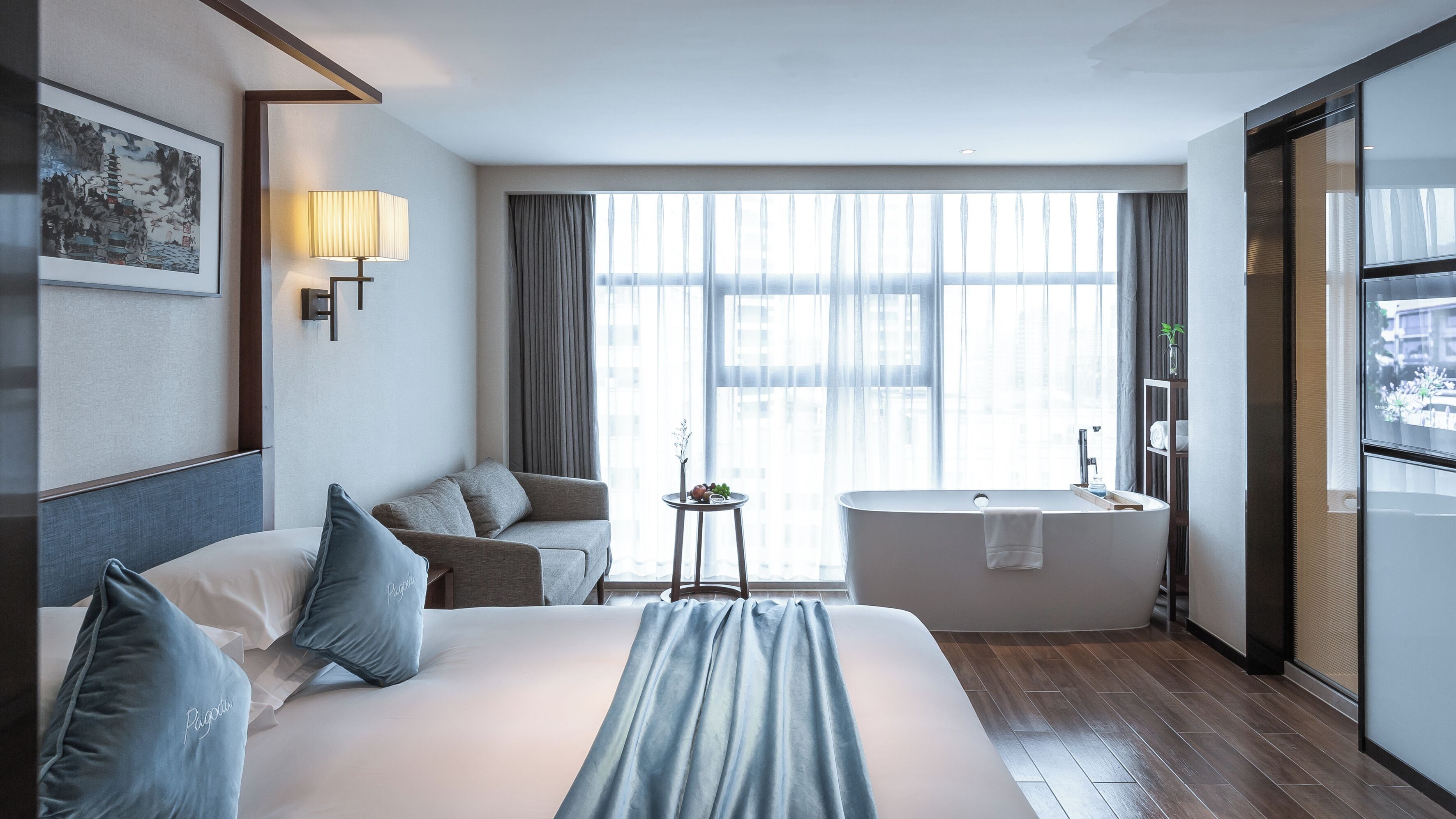 Quarto de hotel com uma cama grande, área de estar com sofá e poltronas, uma pequena mesa com vaso e frutas, banheira e janela com cortinas.