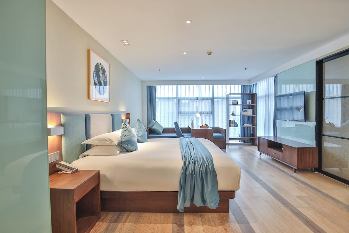 Ein modernes Hotelzimmer mit einem großen Bett, Nachttischen, einem Fernseher, einem Bücherregal und einem großen Fenster mit viel Tageslicht.