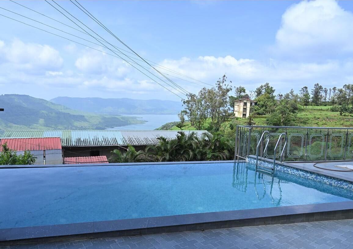 Piscina con vistas a un lago y montañas, rodeada de vegetación y un pequeño edificio.