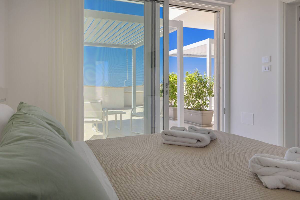 A hotel room with a bed, a glass door leading to an outdoor area with a table and chairs, and a potted plant.