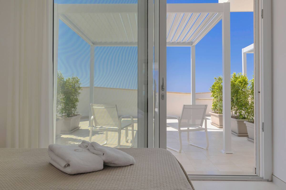 A hotel room with a bed, a set of white towels, and a sliding glass door leading to an outdoor patio with chairs and potted plants.