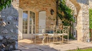 Un bâtiment en pierre avec un toit en tuiles, un patio couvert avec des chaises en métal et un jardin.