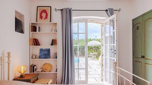 Une chambre avec un lit, une table de chevet avec une lampe, une bibliothèque et une vue sur une terrasse extérieure avec une piscine et des parasols.