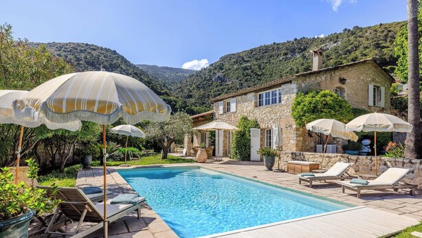Une piscine entourée de bâtiments en pierre et d’une végétation luxuriante, avec des chaises longues et des parasols à proximité.